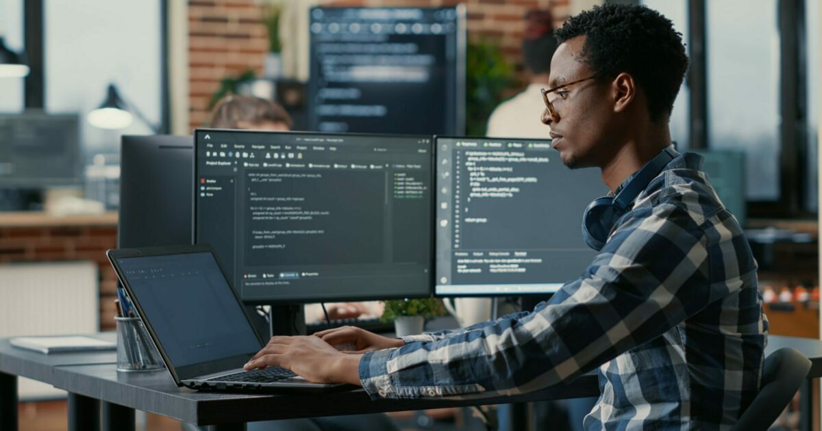 A imagem mostra um desenvolvedor de software trabalhando em um ambiente de escritório moderno. Ele está sentado em frente a três monitores, com dois exibindo códigos de programação. Ele usa um laptop, onde também está programando, e parece concentrado no trabalho. O ambiente é bem iluminado, com uma parede de tijolos ao fundo e algumas plantas decorativas. O desenvolvedor veste uma camisa xadrez azul e branca e usa óculos, com fones de ouvido ao redor do pescoço. (desenvolvedor, programador, código, programação, devs)