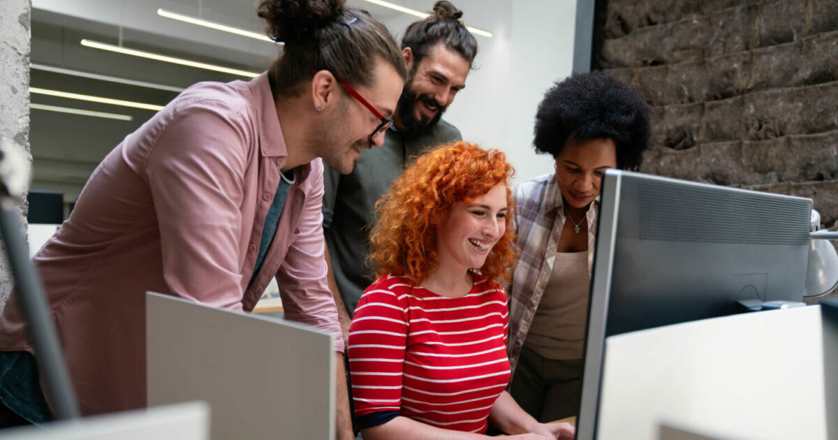 Equipe de trabalho colaborando em um ambiente de escritório moderno. Quatro pessoas, de diferentes perfis, estão reunidas em volta de um computador, sorrindo e discutindo algo com entusiasmo. O ambiente descontraído e criativo reflete o espírito de inovação e trabalho em equipe (desenvolvedores, programadores, time, equipe, trabalho, talentos, talento, Citizen Developer, vagas abertas, aws, campinho digital, soft-skills, cursos, ibm, curso)