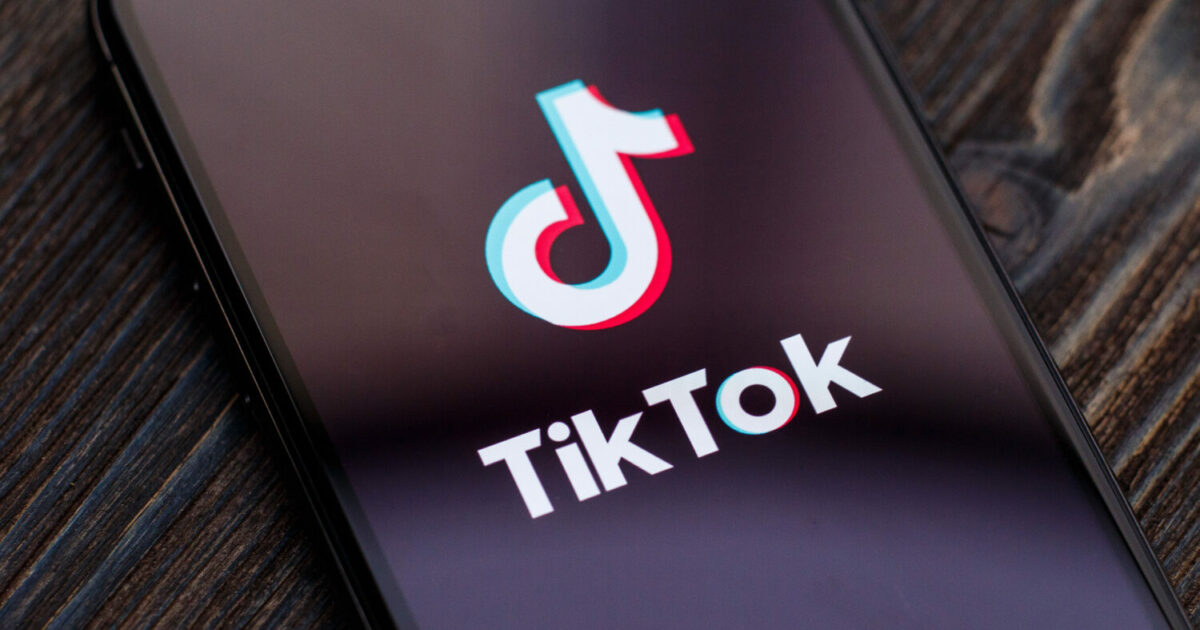 Ícone do aplicativo TikTok exibido na tela de um smartphone sobre uma superfície de madeira escura. O logotipo característico do TikTok aparece com suas cores neon em azul e rosa, contrastando com o fundo preto do dispositivo, representando o uso de redes sociais em dispositivos móveis (Latam, aviões, nebraska)