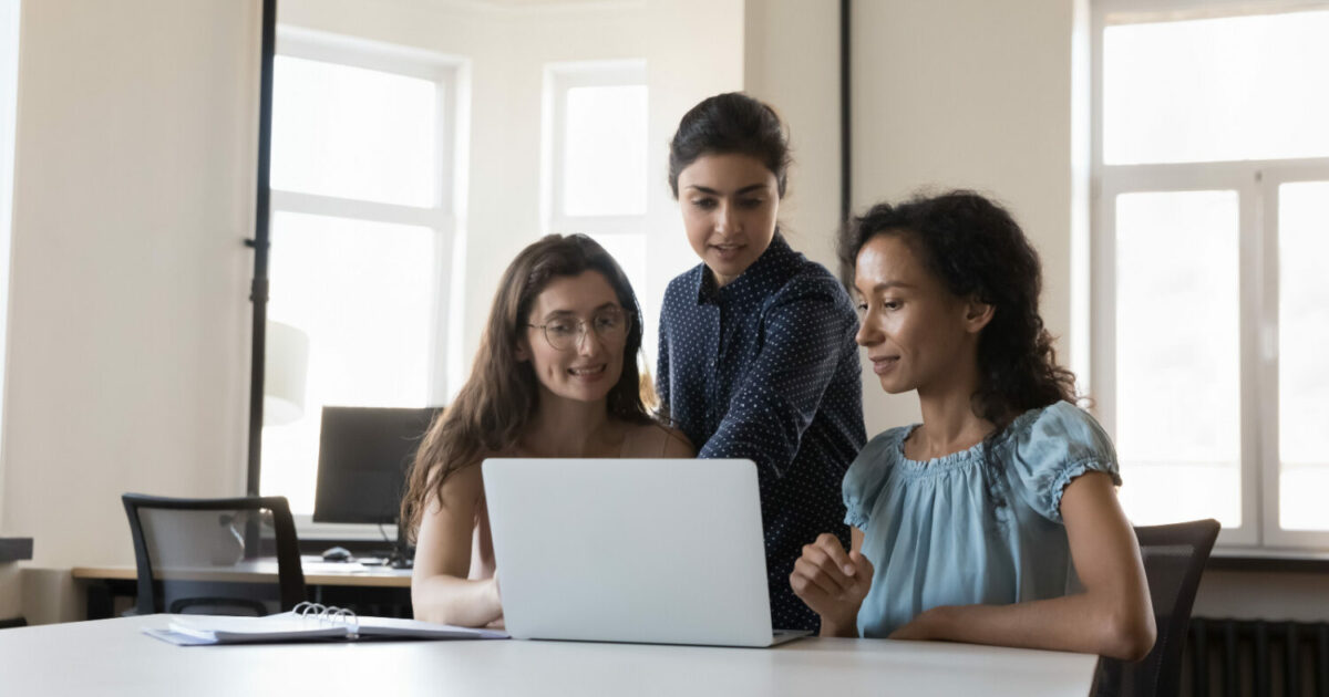 Três mulheres de diferentes origens trabalhando juntas em um escritório. Duas estão sentadas, olhando para a tela de um laptop, enquanto a terceira, de pé, aponta para o computador. O ambiente é iluminado, com janelas amplas ao fundo, simbolizando colaboração e diversidade no trabalho (mulheres, TI, instituto, tecnologia, diversidade, inclusão, mercado de TI, vagas de diversidade, startups brasileiras, huawei, MEC)