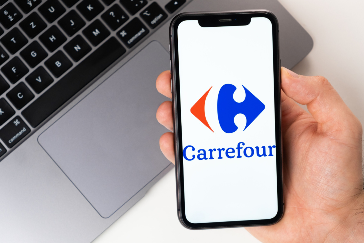 A imagem mostra uma mão segurando um smartphone, que exibe o logotipo do Carrefour na tela. O logo consiste em um ícone azul e vermelho ao lado do nome "Carrefour" escrito em azul. Ao fundo, há um laptop prateado com teclado preto, parcialmente visível. A cena sugere um ambiente de trabalho ou um contexto digital relacionado à marca Carrefour.