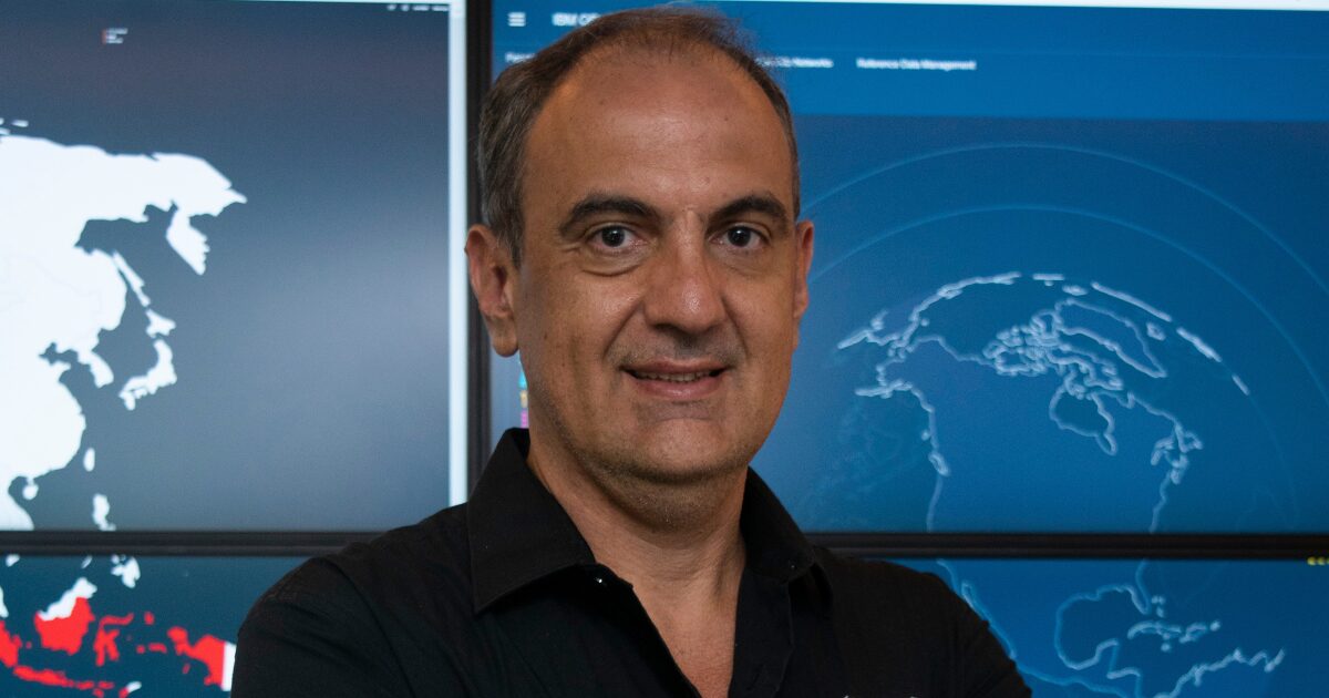 Marcelo Branquinho, CEO da TI Safe, posando com os braços cruzados em frente a monitores exibindo mapas e dados digitais. Ele usa camisa preta e tem expressão confiante, representando liderança no setor de cibersegurança