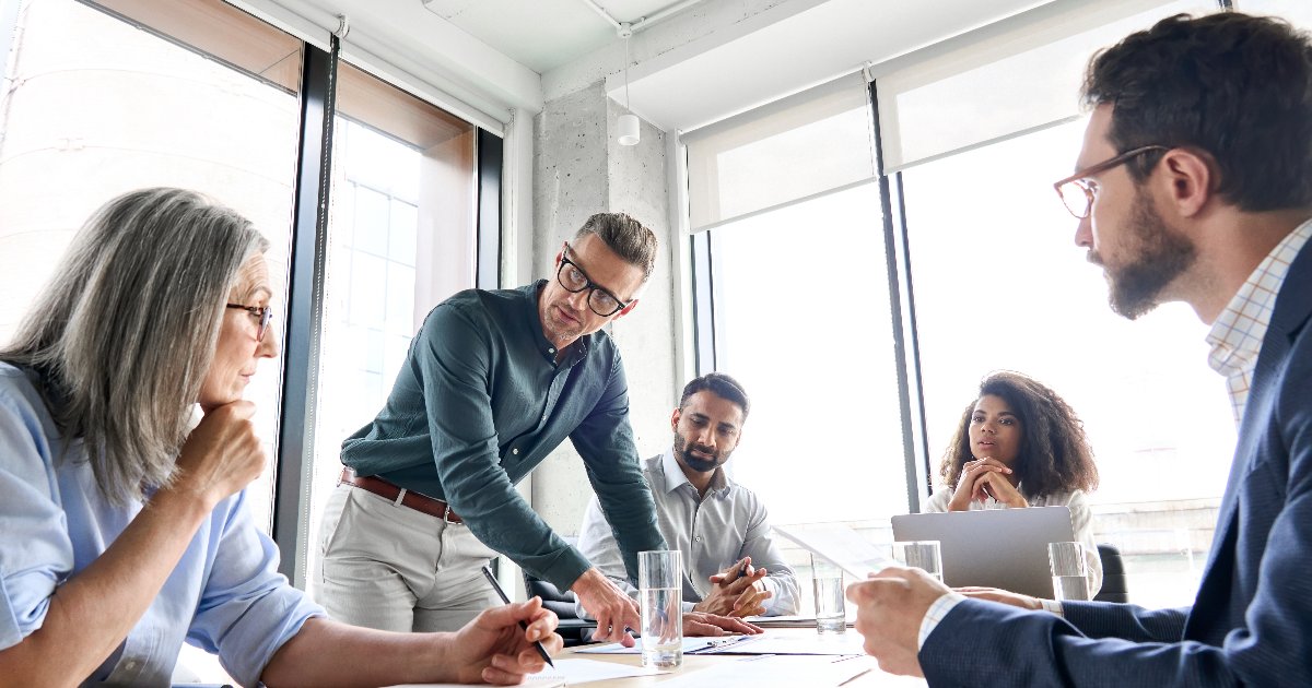 Grupo de profissionais em uma reunião de trabalho em um ambiente iluminado por luz natural. Um homem de óculos, em pé, aponta para documentos sobre a mesa enquanto os outros membros da equipe, sentados, o escutam atentamente. A equipe é diversa, com diferentes idades e etnias, refletindo colaboração e discussão em um ambiente corporativo moderno (vagas, oportunidade, reunião, executivos, encontro, negócios, liderança, abordagens, empresas familiares, líder, propósito, reuniões)