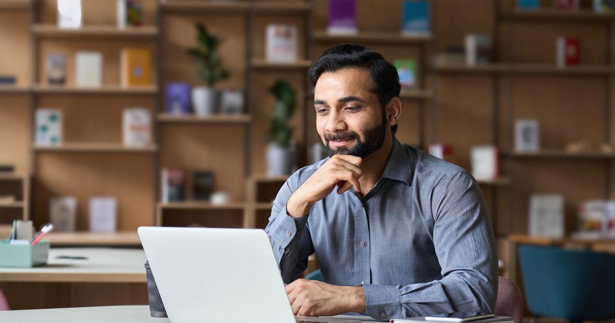 Homem de camisa social cinza sentado em frente a um laptop branco, sorrindo levemente enquanto olha para a tela. Ele está em um ambiente interno moderno, possivelmente uma biblioteca ou espaço de trabalho compartilhado, com prateleiras de madeira ao fundo contendo livros, vasos de plantas e objetos decorativos. (oportunidade em ti, executivo, vagas, cursos, empresas