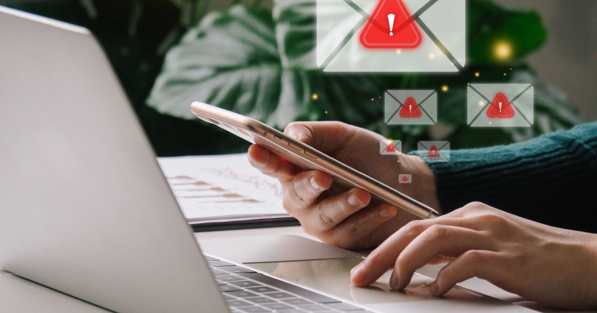 Pessoa segurando um celular enquanto utiliza um notebook, com ícones de e-mails flutuando na tela, todos com um símbolo de alerta vermelho, representando mensagens de spam, scam ou phishing (golpes virtuais). Ao fundo, uma planta desfocada. (e-mail, spam, scam, phishing, golpe, check point, américa do sul, governo)