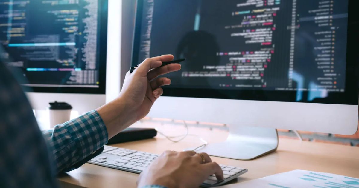 Imagem de um desenvolvedor de software trabalhando em frente a dois monitores grandes exibindo códigos de programação. A pessoa está com uma camisa xadrez azul, segurando uma caneta enquanto interage com o teclado. A cena retrata um ambiente de trabalho moderno e focado em tecnologia, com códigos detalhados na tela e itens de escritório ao fundo (desenvolvedor, programador, empresas, profissional, trabalhador, código, programação, PageGroup)