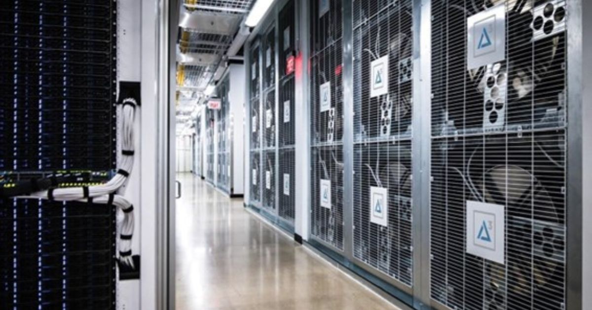 A imagem mostra o interior de um data center moderno, com corredores alinhados por servidores e equipamentos de armazenamento de dados. No lado esquerdo, há racks de servidores com cabos organizados e luzes indicadoras de funcionamento. No lado direito, grandes estruturas metálicas com grades protegem equipamentos de resfriamento e processamento, exibindo logotipos. O ambiente é bem iluminado e possui um piso refletivo, transmitindo uma atmosfera tecnológica e de alta segurança, essencial para a infraestrutura de armazenamento e processamento de dados em larga escala (odata, data center, méxico)