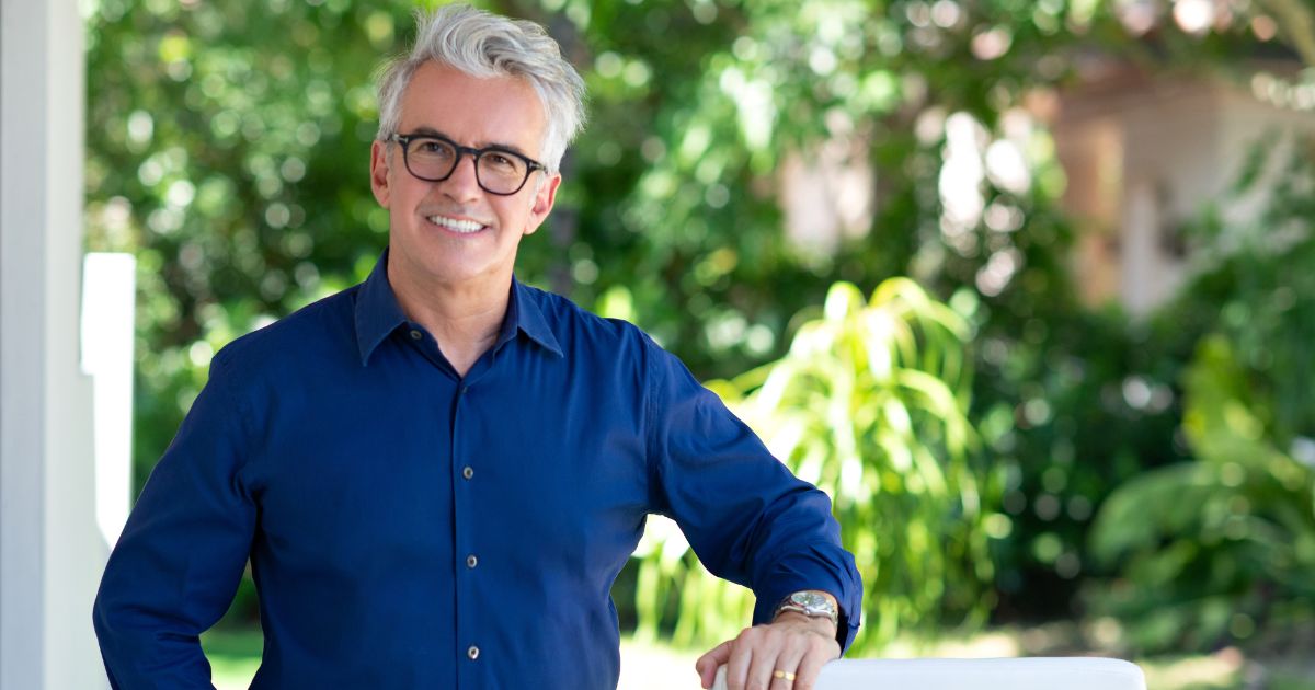Gilmar Batistela, CEO da Evox, sorrindo em um ambiente externo rodeado por plantas verdes. Ele está vestindo uma camisa azul escura, com os braços cruzados sobre uma superfície branca. Gilmar tem cabelos grisalhos e usa óculos, exibindo uma expressão amigável e confiante.