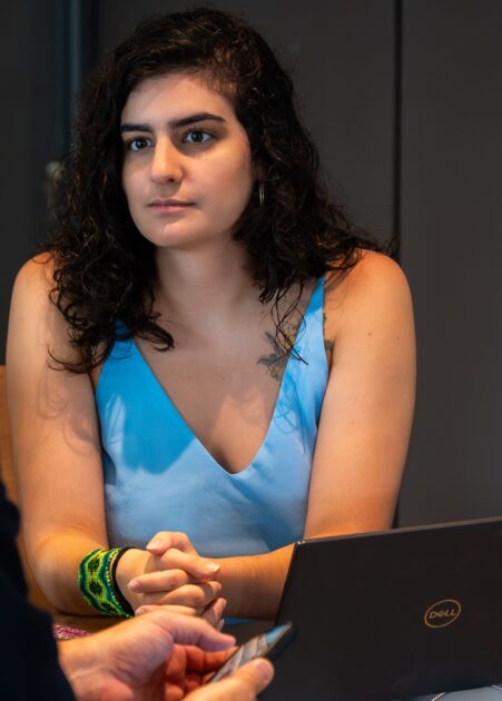 Uma mulher de cabelos encaracolados escuros, vestida com um top azul claro, está sentada à frente de um portátil da marca Dell. Ela tem uma expressão atenta e está com as mãos cruzadas, como se estivesse a ouvir ou participar numa conversa. Usa uma pulseira de missangas verde e preta no pulso esquerdo e tem uma tatuagem visível no ombro. Em primeiro plano, vê-se parcialmente a mão de outra pessoa a segurar um telemóvel. O ambiente parece ser de uma reunião informal ou entrevista.