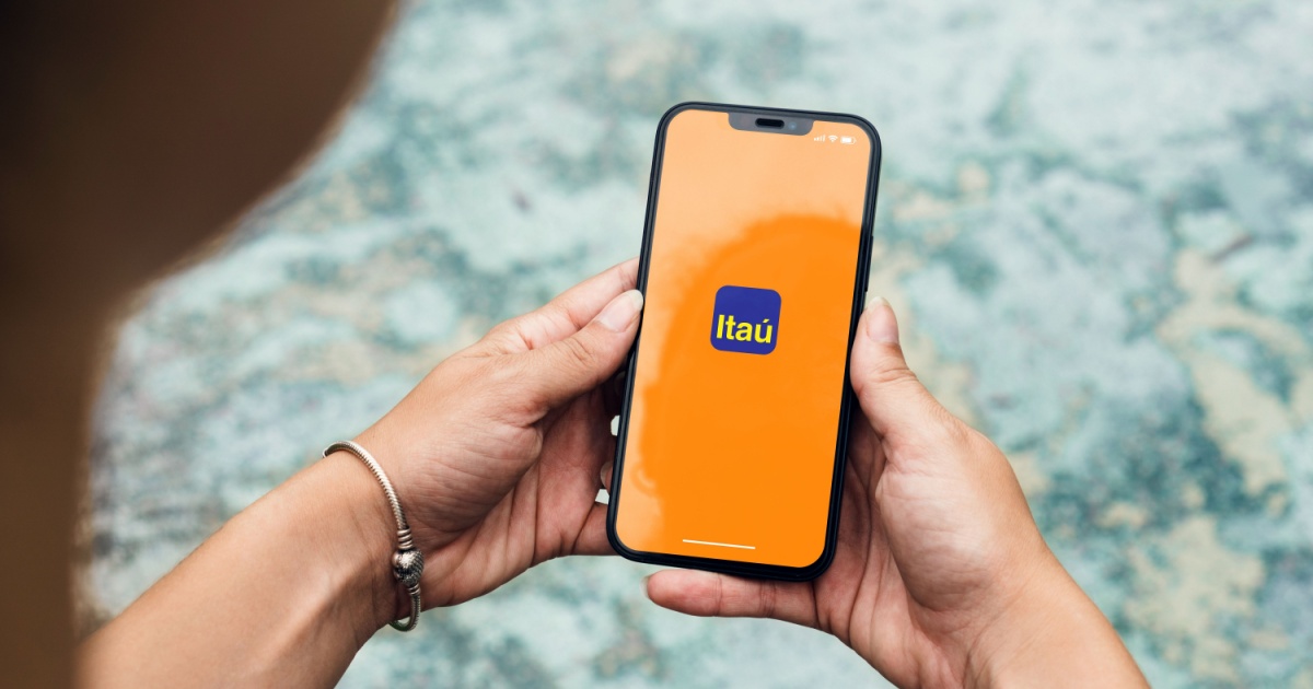 Pessoa segurando um smartphone com a tela exibindo o logotipo do Itaú sobre um fundo laranja. A imagem destaca o uso do aplicativo bancário, simbolizando praticidade e tecnologia, com um fundo desfocado que dá foco à interação com o dispositivo (itau, itau unibanco, app, superapp, inteligencia itau)