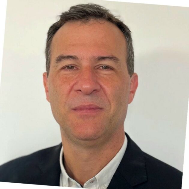 Um homem de pele clara, cabelo curto e escuro, usando um blazer preto e camisa branca, posando para uma foto. O fundo é neutro, branco, e a imagem parece ser um retrato profissional.