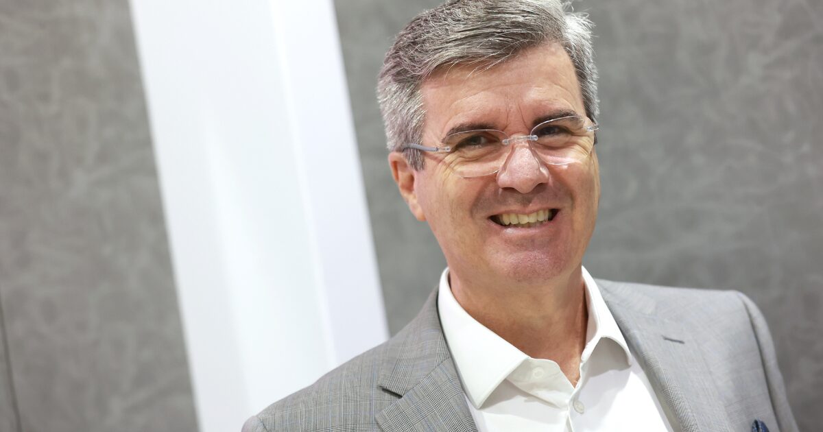 A imagem mostra um homem de óculos e cabelo grisalho, sorrindo para a câmara. Ele veste um fato cinzento claro e uma camisa branca, transmitindo uma aparência profissional e amigável. O fundo é neutro, com tons de cinza e branco, destacando o sujeito na fotografia. A iluminação é suave, criando um ambiente limpo e corporativo.