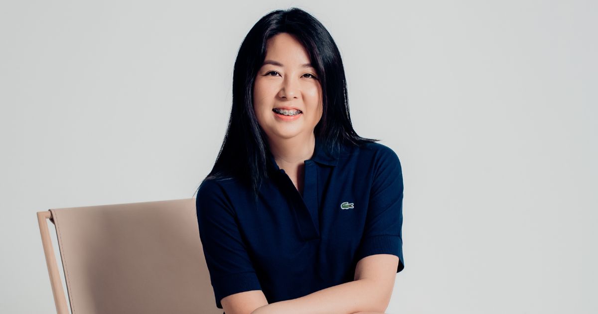 "Debora Noemi, diretora de produtos e sócia da Happy, sentada em uma cadeira bege, vestindo uma camisa polo azul escuro com o logotipo da Lacoste. Ela está sorrindo e tem cabelos longos e escuros. Foto: Divulgação."