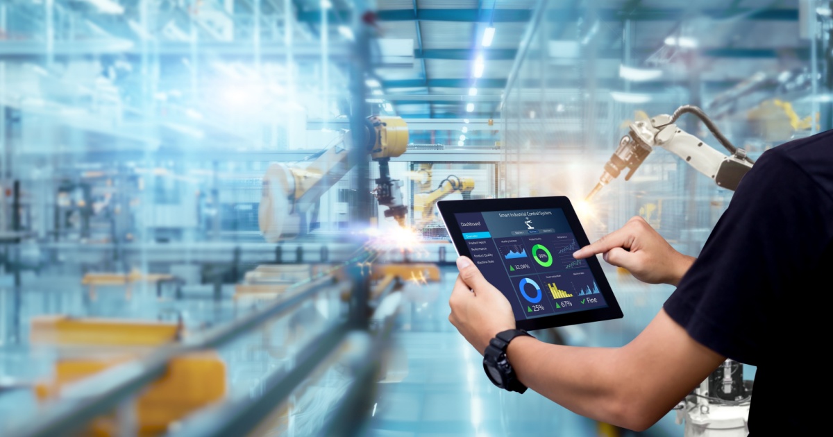 Um ambiente industrial moderno com braços robóticos automatizados ao fundo. Em destaque, uma pessoa segurando um tablet que exibe gráficos e informações sobre o sistema de controle da produção, representando conceitos de automação e Indústria 4.0. O ambiente é iluminado e possui linhas de montagem visíveis ao longo da fábrica (indústria, automação, robos, indústria 4.0, GS1, dados, futuro)