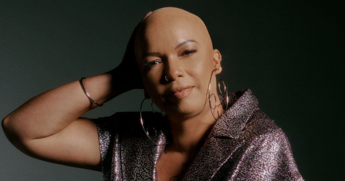 Fotografia de Grazi Mendes, head de Diversidade, Equidade e Inclusão da ThoughtWorks. Ela aparece em um retrato expressivo, com a cabeça raspada, usando argolas grandes e um look brilhante em tons metálicos. Seu olhar é confiante e sereno, com um leve sorriso, enquanto apoia uma das mãos na cabeça. O fundo é escuro, destacando sua presença e estilo sofisticado.