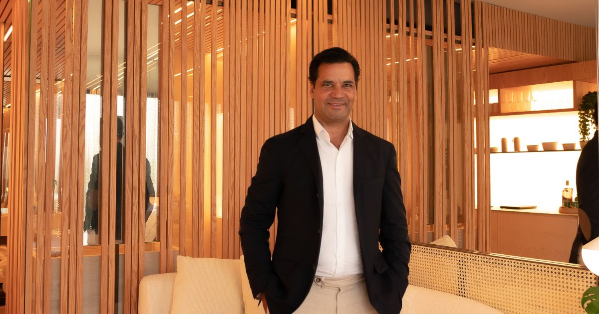 Gustavo Filgueiras, CEO do Grupo Emiliano, posa para a foto com um sorriso, vestindo um blazer preto sobre uma camisa branca e calça bege. O ambiente sofisticado e bem iluminado apresenta painéis de madeira verticalizados, decoração minimalista e tons neutros, transmitindo elegância e modernidade.
