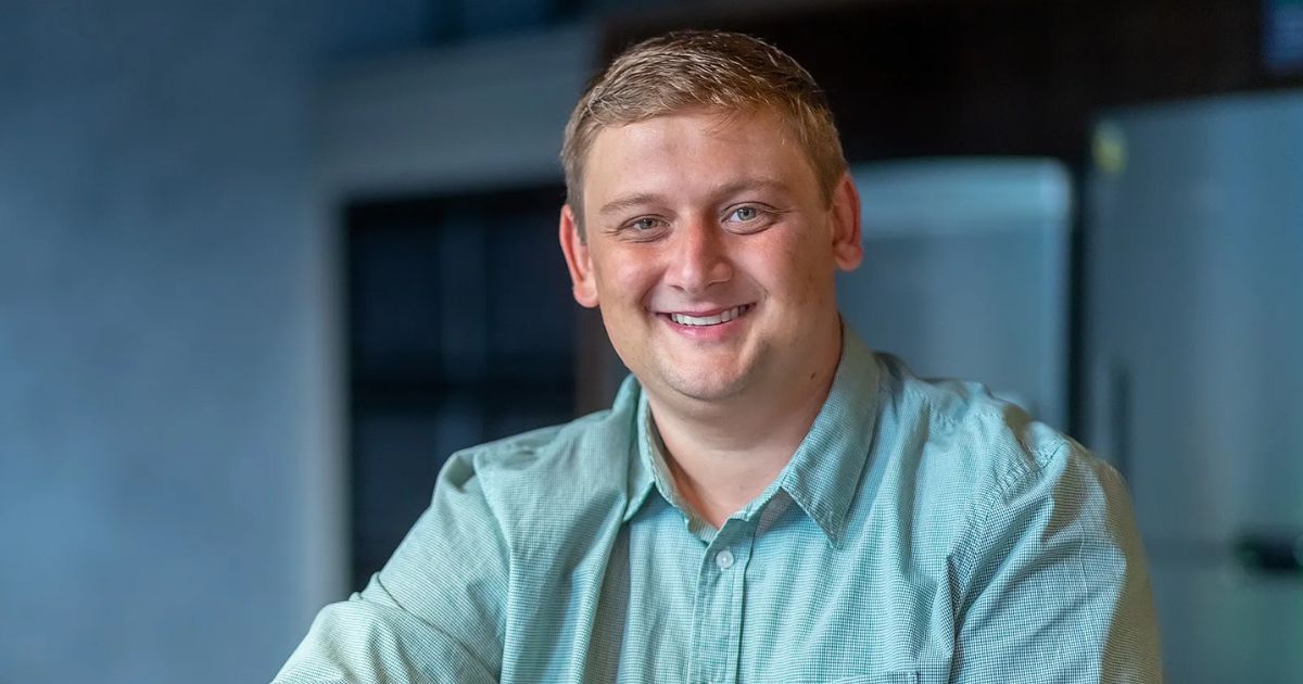 Gustavo Salles, CEO e fundador da SalesHunter, sorrindo para a câmera. Ele veste uma camisa verde e está em um ambiente interno com fundo desfocado, que inclui uma geladeira e janelas escuras (Gustavo Salles, SalesHunter, CRO)