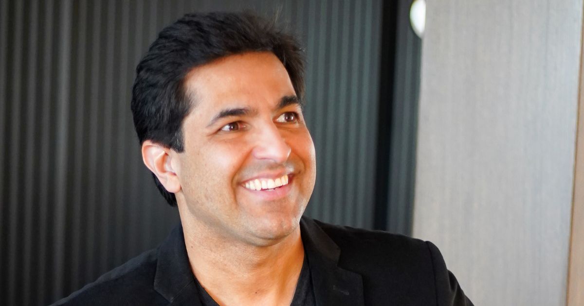 Um homem de pele morena e cabelos escuros sorri amplamente enquanto olha para o lado. Ele veste um blazer preto sobre uma camisa escura, transmitindo uma imagem profissional e sofisticada. O fundo tem um tom moderno, com elementos em preto e madeira, criando um ambiente elegante e contemporâneo (CEO, Prime Control, Marcio Viana)