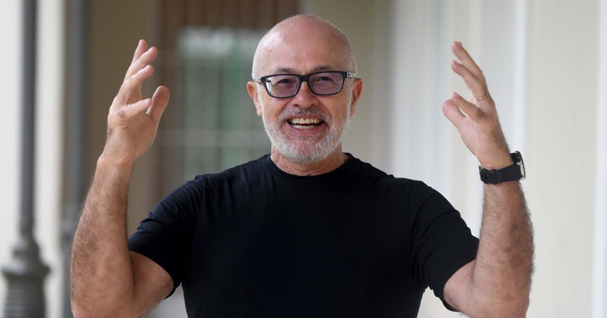 Homem de barba branca e óculos de armação preta sorriem extensamente enquanto gesticula com as mãos erguidas. Ele veste uma camisa preta e um relógio no pulso esquerdo. O fundo da imagem está desfocado, indicando um ambiente ao ar livre ou um corredor iluminado. Trata-se de Silvio Meira, fundador do CESAR , referência em inovação e tecnologia no Brasil.