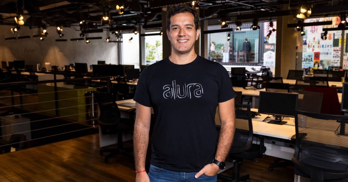 Adriano Almeida, novo CEO da Alura, sorrindo e posando em pé, usando uma camiseta preta com o logotipo "Alura", em um ambiente moderno de escritório com várias estações de trabalho ao fundo.