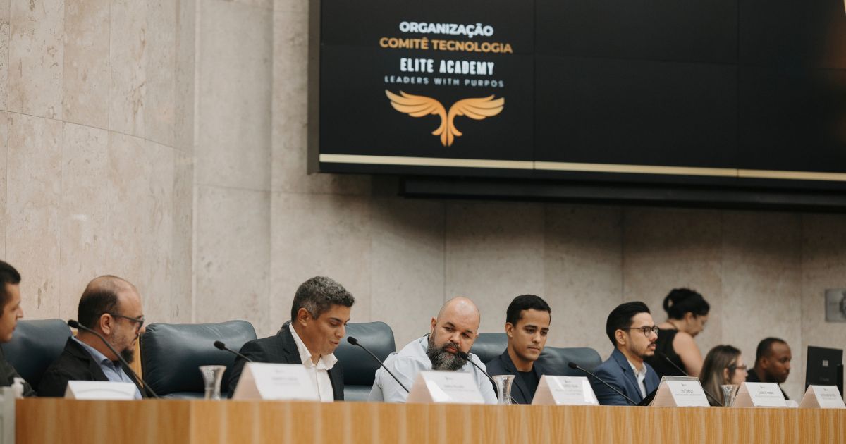 Painel de discussão com sete homens sentados lado a lado em uma mesa de madeira clara, em um auditório com parede de mármore. Ao fundo, um telão exibe o logotipo da Elite Academy, com a frase “Organização Comitê Tecnologia – Elite Academy – Leaders with Purpose” e um ícone dourado em forma de asas estilizadas. Todos estão com microfones à frente e placas de identificação sobre a mesa. Algumas pessoas aparecem ao fundo, em pé