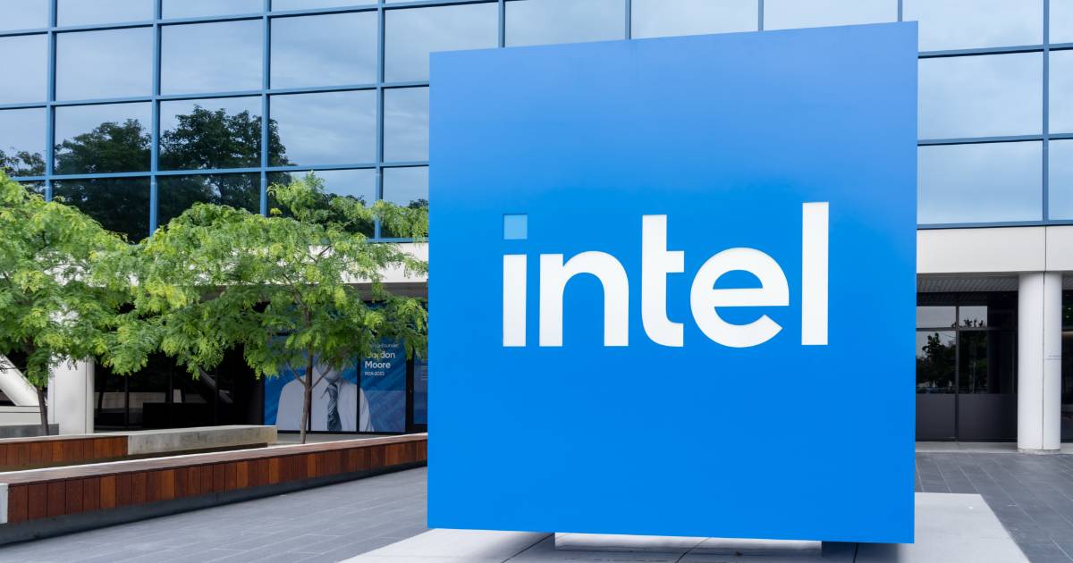 A imagem mostra um grande letreiro azul com o logotipo da Intel em branco, localizado na parte externa de um prédio moderno com fachada de vidro espelhado. Árvores e bancos de madeira compõem a paisagem ao redor da entrada. A cena reflete um ambiente corporativo e tecnológico, chips