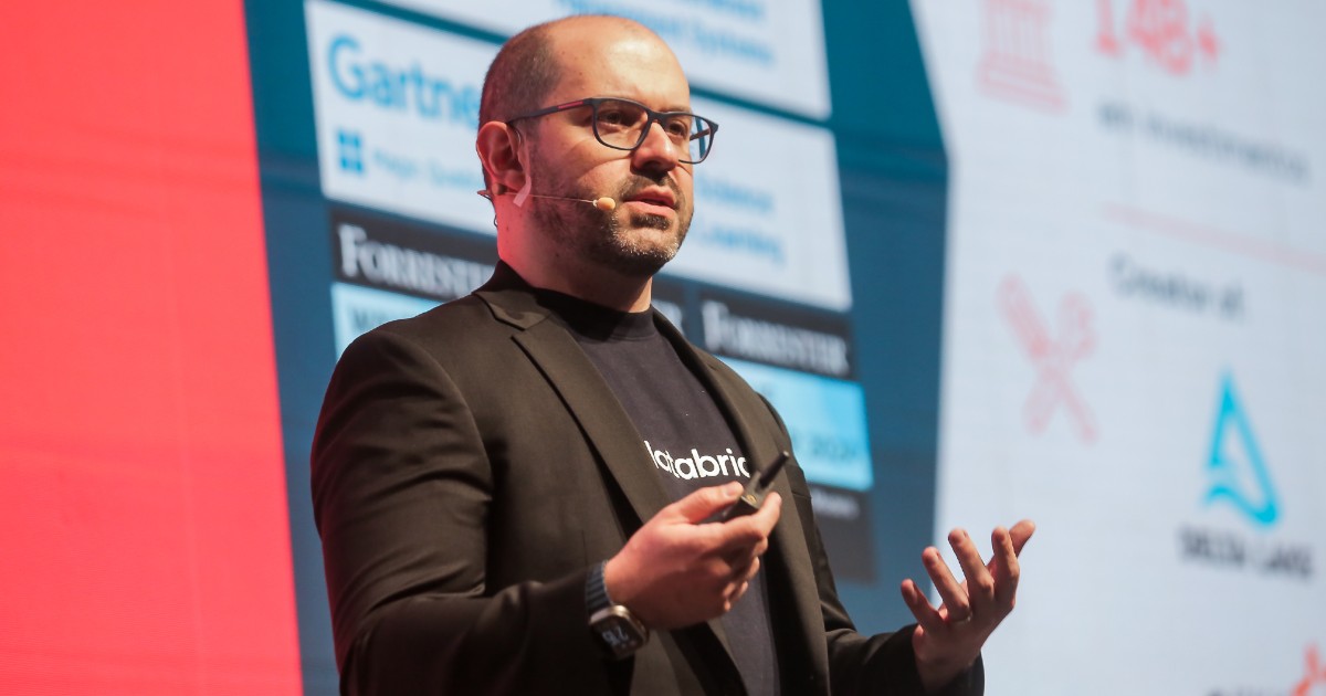 Marcos Grilanda, vice-presidente e gerente geral da Databricks para a América Latina, aparece no palco durante o evento Data Intelligence Day da Databricks. Ele está em pé ao lado de um púlpito transparente com o logotipo da empresa, vestindo blazer preto e camiseta preta com a marca Databricks. Ao fundo, há um grande telão vermelho e azul exibindo dados de crescimento da empresa na região LATAM, destacando um aumento de 82% em 2024 e 66 mil usuários diários