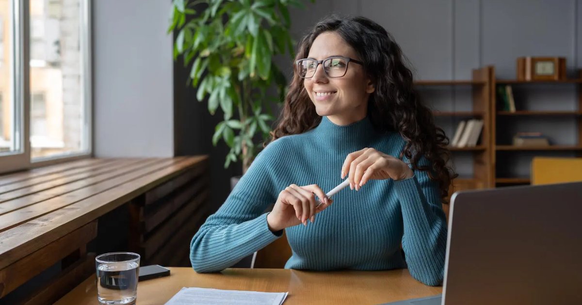 Mulher sorridente sentada à mesa de trabalho em um ambiente moderno e bem iluminado, olhando pela janela com expressão de satisfação. Ela usa óculos e um suéter azul, segura uma caneta e tem à sua frente um copo de água, papéis e um notebook. Ao fundo, há uma estante com livros e uma planta, transmitindo uma atmosfera de tranquilidade e produtividade, associada à satisfação e bem-estar no trabalho. (mulher, trabalho, satisfação, felicidade, alegria)