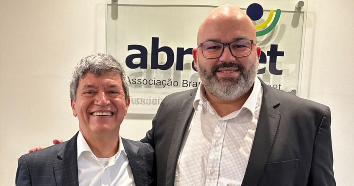 Dois homens de terno estão sorrindo e posando para a foto em frente a uma placa da "Abranet" (Associação Brasileira de Internet). Um dos homens tem cabelo grisalho e está vestindo um terno escuro, enquanto o outro tem barba, usa óculos e veste um terno preto com uma camisa clara.