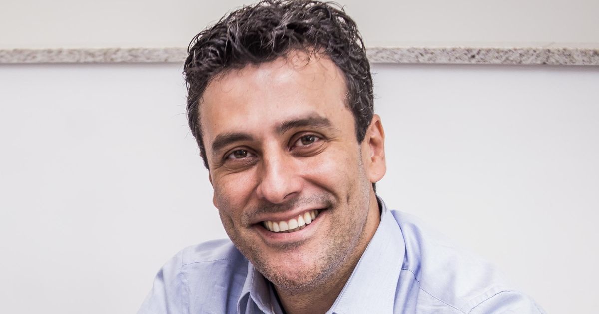 Homem sorridente com cabelo escuro e encaracolado, vestindo camisa azul clara, posando diante de uma parede branca com uma superfície de granito ao fundo. A expressão facial transmite simpatia e cordialidade.