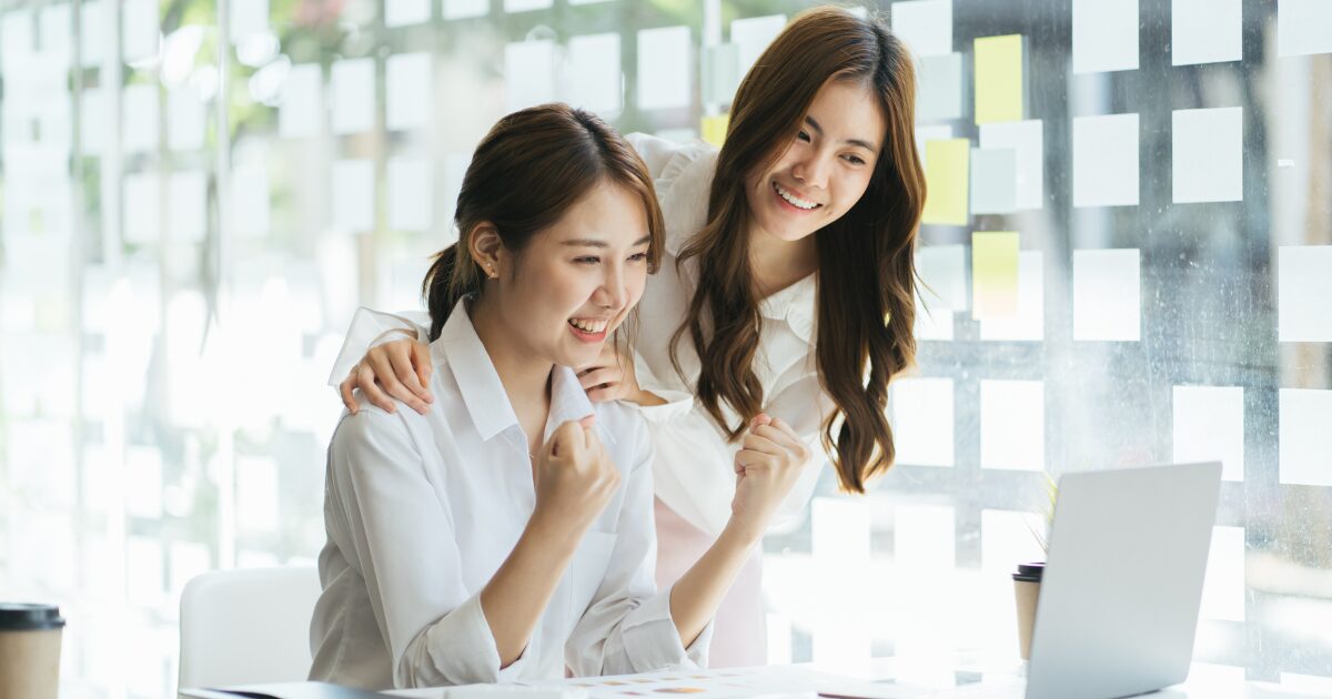 Duas mulheres felizes comemoram juntas em frente a um portátil, num ambiente de escritório moderno com luz natural e notas adesivas coladas no vidro.