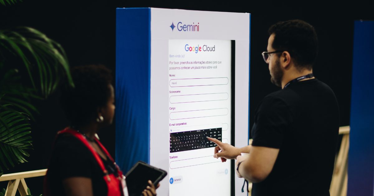 Fotografia de duas pessoas interagindo com um quiosque digital da Google Cloud, com a marca Gemini no topo. Um homem aponta para o ecrã tátil onde aparece um formulário de preenchimento de dados, enquanto uma mulher observa segurando um tablet. O ambiente é interno, com iluminação suave e algumas plantas decorativas ao fundo.