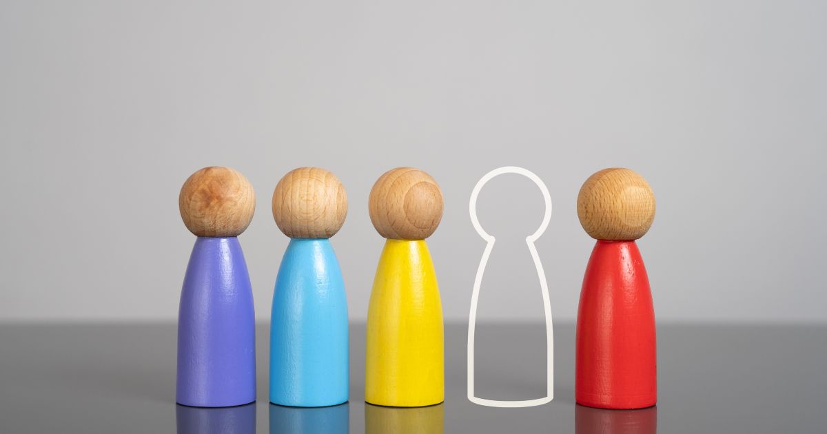 Cinco figuras de madeira com cabeças esféricas e corpos pintados de cores diferentes (roxo, azul claro, amarelo, espaço vazio com contorno branco, e vermelho) alinhadas em fila. O fundo é cinzento claro e a superfície é refletora.