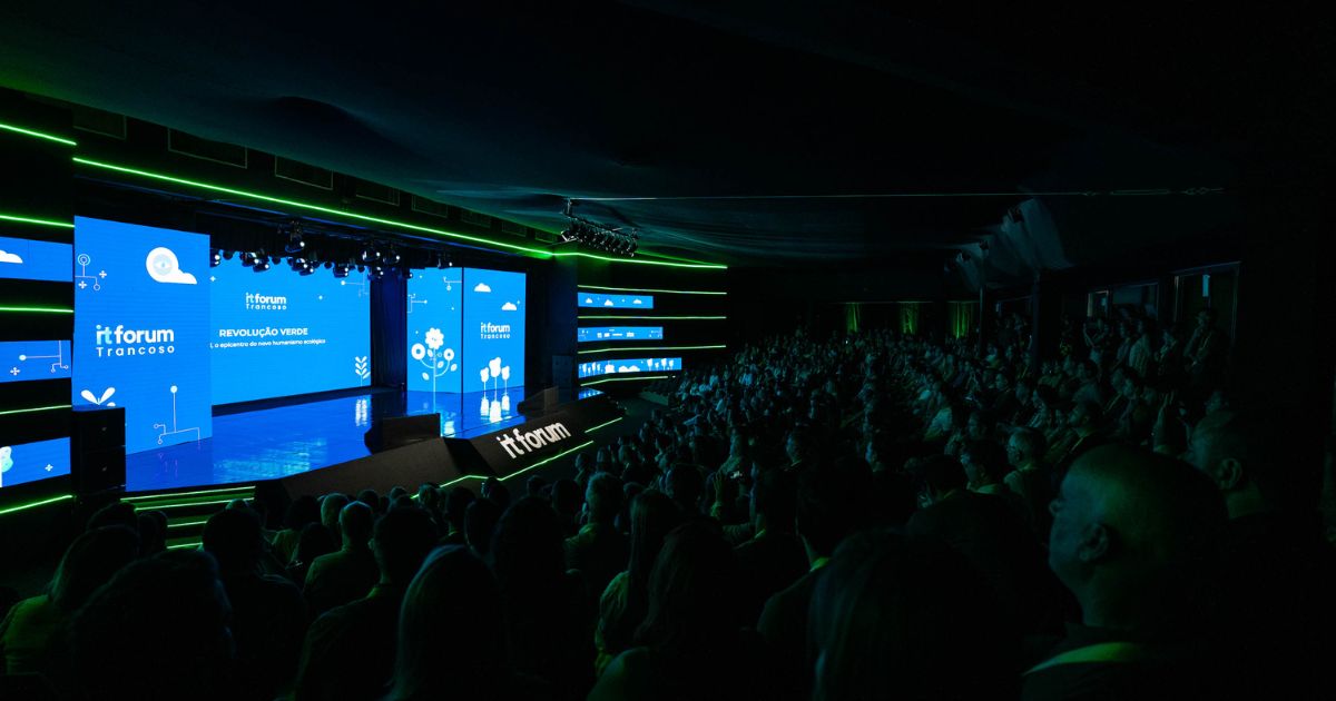 Fotografia de uma grande conferência tecnológica em auditório escuro, com iluminação em tons de verde e azul. O palco exibe o nome 'itforum Trancoso' em ecrãs digitais com gráficos relacionados com sustentabilidade e tecnologia, incluindo plantas e ícones de nuvens. O tema destacado é 'Revolução Verde', com o subtítulo 'A experiência do novo humanismo ecológico'. A plateia está cheia, com o público atento à apresentação.