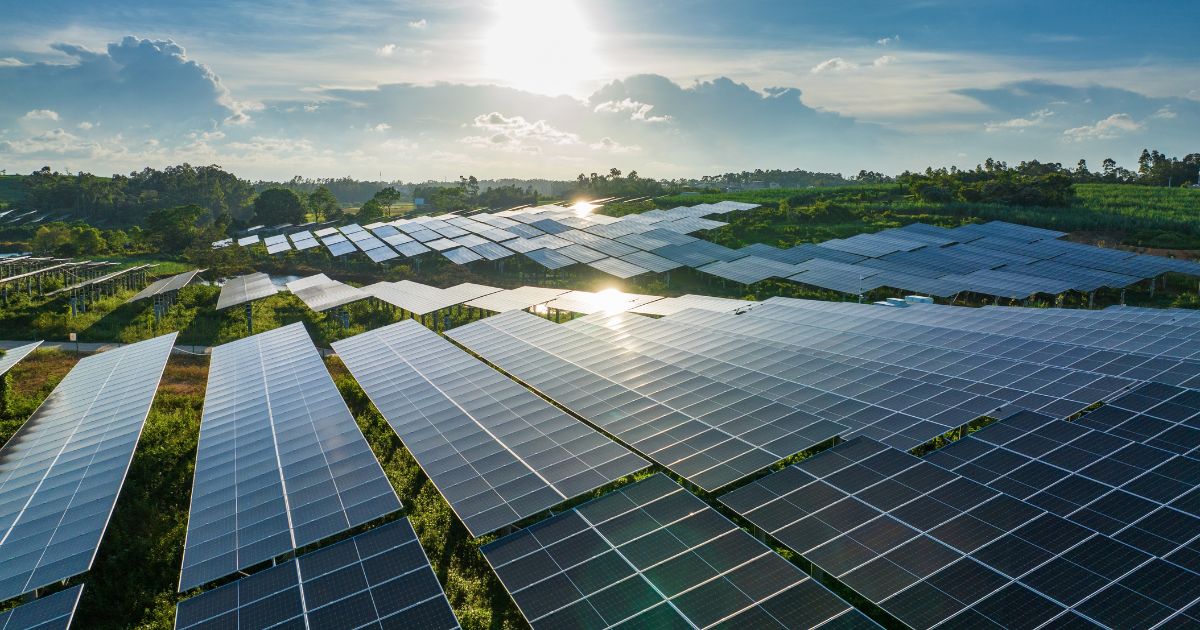 Extensa fazenda solar com múltiplos painéis fotovoltaicos refletindo a luz do sol ao entardecer. O cenário é rodeado por vegetação e colinas ao fundo, sob um céu azul com algumas nuvens. A imagem transmite a ideia de energia renovável e sustentabilidade (desafios regulatórios)