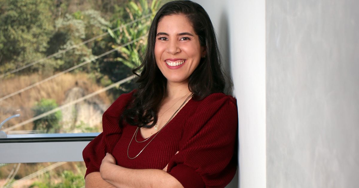 Uma mulher jovem, de cabelo comprido e escuro, está encostada a uma parede branca com os braços cruzados e um largo sorriso. Ela usa uma blusa de manga comprida cor de vinho e um colar delicado. Atrás dela, através de uma janela, vê-se vegetação e fios de eletricidade, indicando um ambiente urbano próximo à natureza.