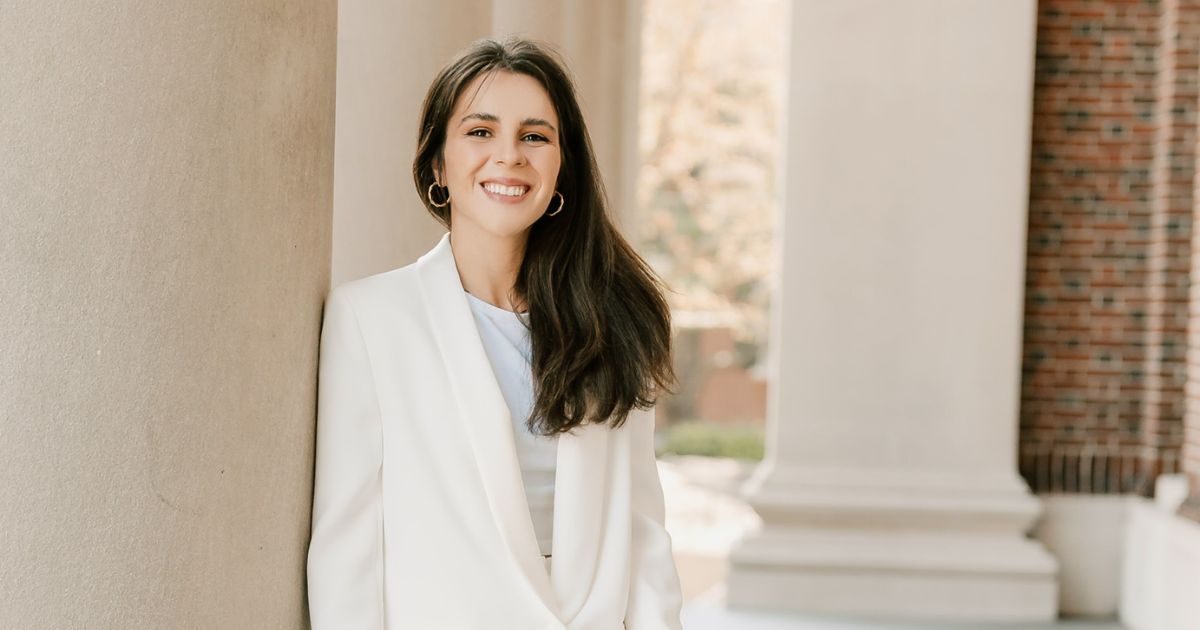 Mulher jovem a sorrir, vestida com blazer branco, encostada a uma coluna de pedra num espaço exterior com arquitetura clássica ao fundo.