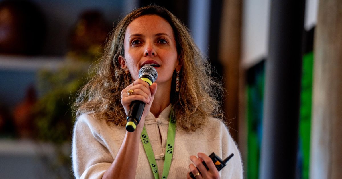 Mulher jovem com cabelo comprido e ondulado, a falar ao microfone durante uma apresentação. Está vestida com uma camisola clara e também tem um crachá verde ao pescoço. Segura um microfone na mão direita e um comando ou dispositivo na esquerda, com fundo interior desfocado (regeneração, esg, economia regenerativa)