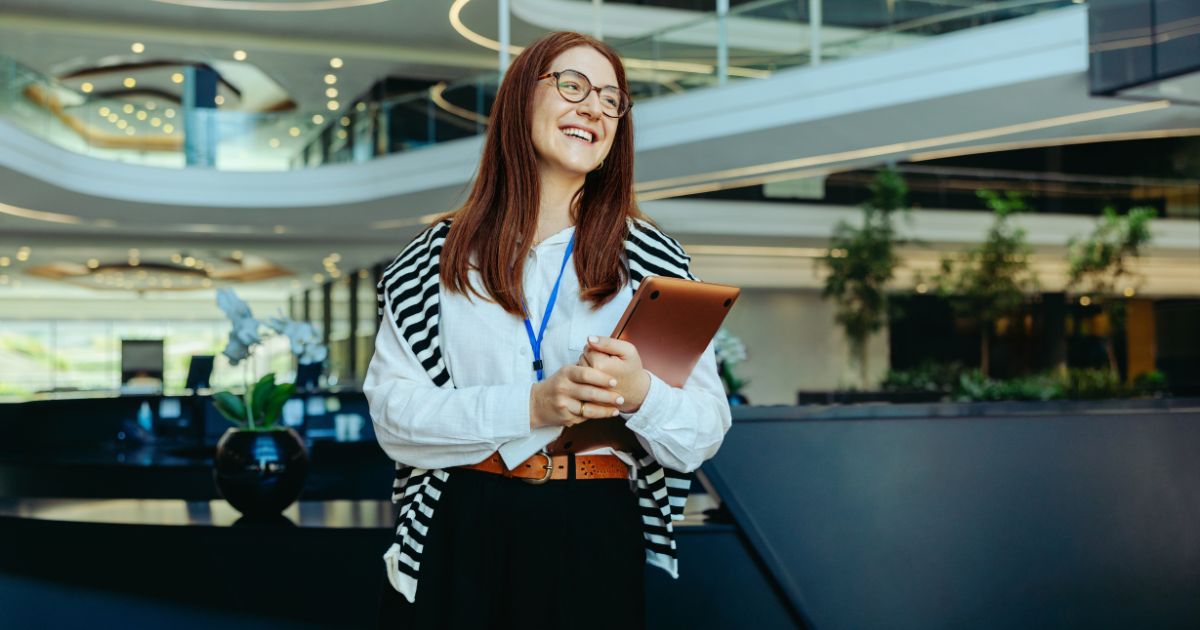 Mulher jovem com cabelos ruivos e óculos sorrindo enquanto segura um tablet em um ambiente corporativo moderno. Ela veste uma camisa branca, um cinto marrom e tem um crachá azul pendurado no pescoço. Um suéter listrado preto e branco está apoiado sobre os ombros. O fundo mostra um espaço amplo e sofisticado, com plantas, iluminação elegante e arquitetura contemporânea.