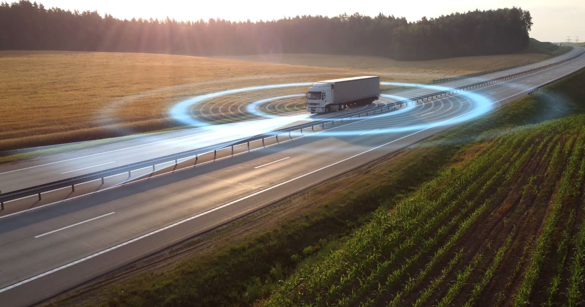 Um caminhão branco trafega em uma rodovia cercada por campos verdes e dourados ao pôr do sol. Há um efeito gráfico azul simulando ondas ao redor do veículo, sugerindo tecnologia de sensores ou conectividade digital na estrada (cimento)