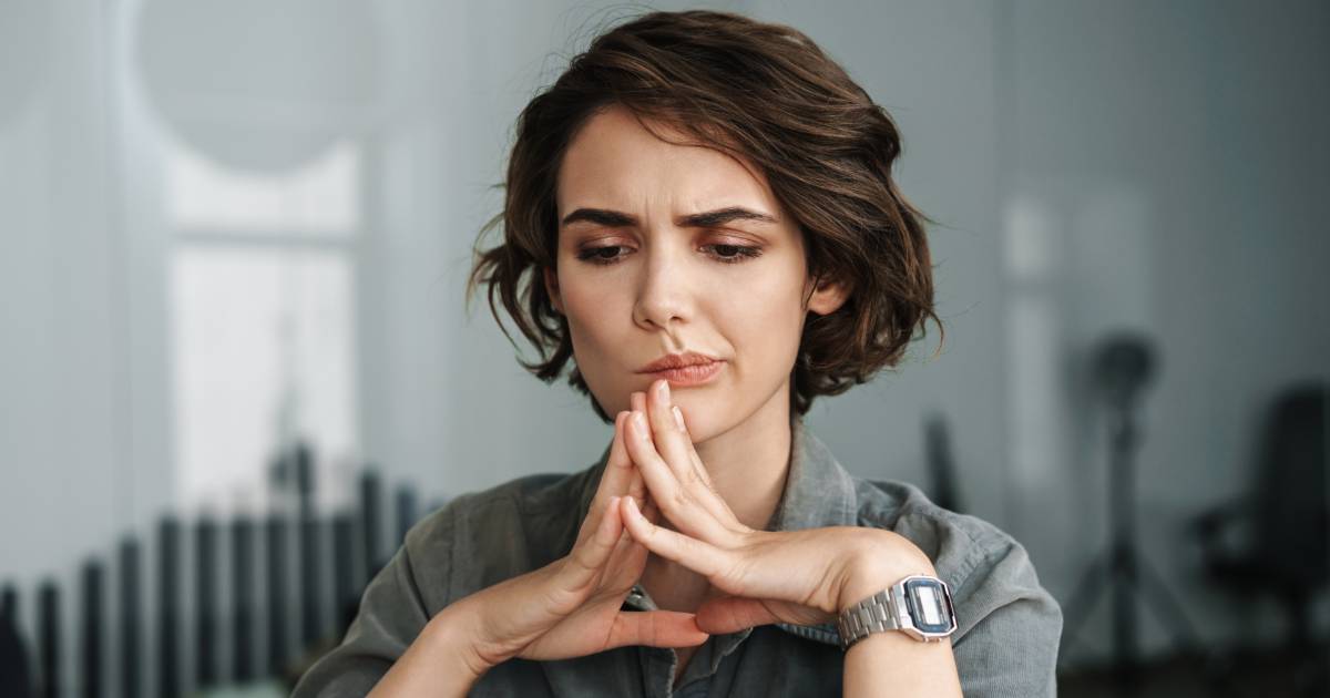 Mulher de cabelos castanhos curtos e camisa cinza aparece pensativa, com as mãos unidas em frente ao rosto, em um ambiente interno desfocado, sugerindo concentração ou tomada de decisão. Ela usa um relógio metálico no pulso esquerdo (ano)