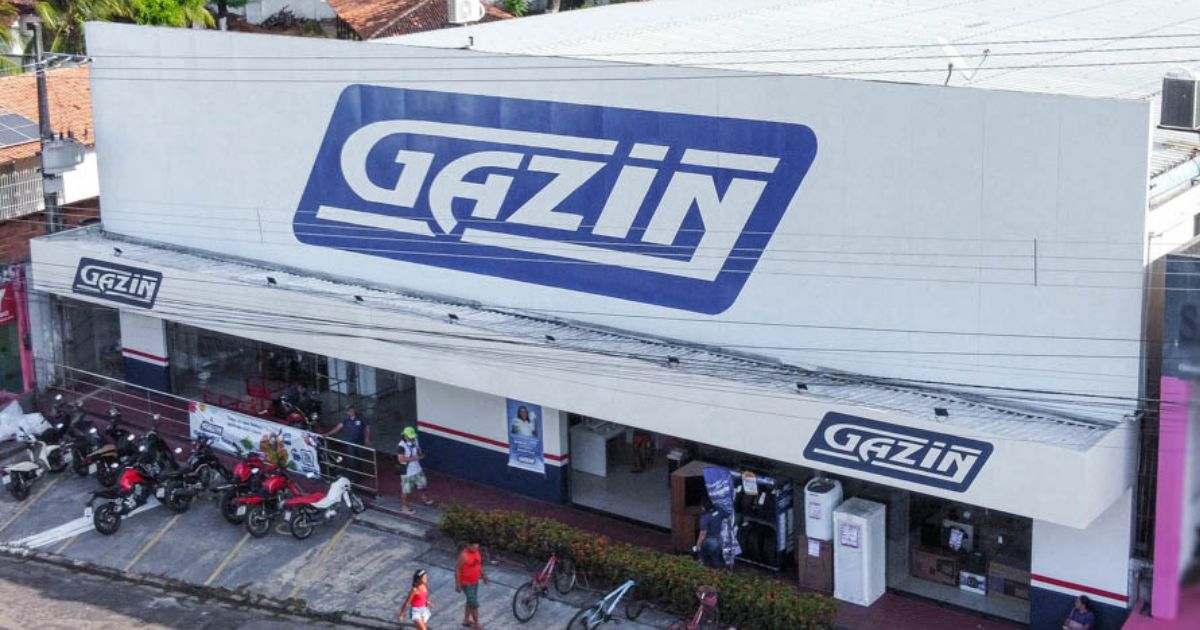 Fachada de uma loja da rede Gazin, com grande letreiro azul e branco exibindo o nome da marca. Na frente do estabelecimento, há diversas motocicletas estacionadas e algumas pessoas caminhando na calçada. A entrada da loja possui vitrines e produtos expostos, sugerindo um ambiente comercial movimentado