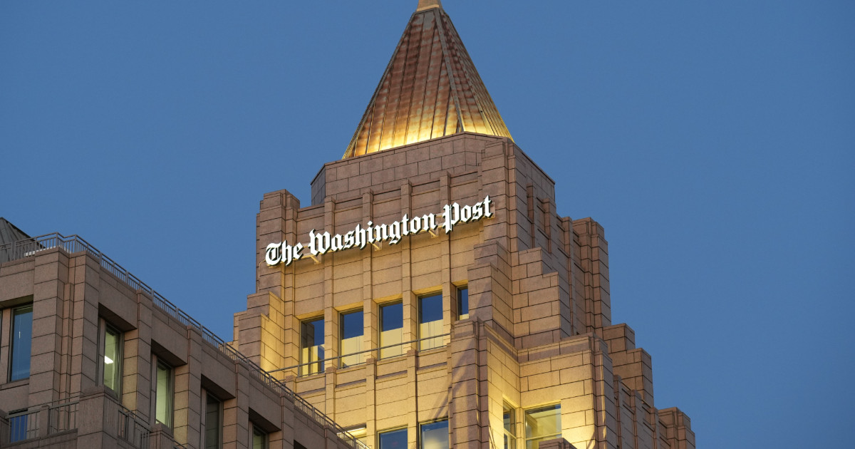 Imagem da parte superior do edifício do jornal "The Washington Post", com destaque para o nome da empresa iluminado em letras brancas. A arquitetura do prédio apresenta um estilo moderno com linhas geométricas e um topo em formato de pirâmide revestido por painéis metálicos dourados. O céu ao fundo está limpo e azul, indicando que a foto foi tirada ao entardecer (direitos)