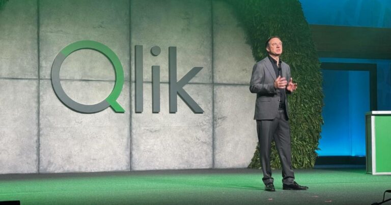 Mike Capone, CEO da Qlik, está posando em frente a um grande letreiro com as letras estilizadas da palavra "Qlik", em um ambiente interno com cortina preta ao fundo. Ele veste um terno escuro com camiseta preta e tem expressão séria.