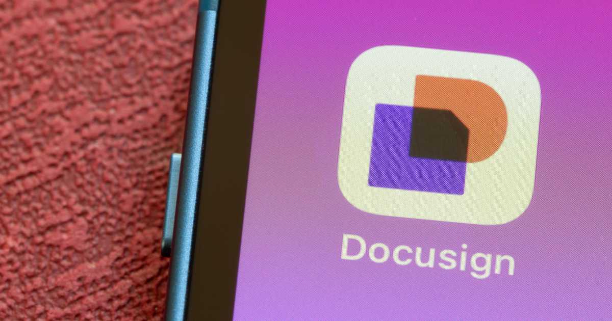 Imagem mostra a tela de um smartphone com o ícone do aplicativo DocuSign em destaque. O ícone tem fundo branco com formas geométricas nas cores roxa, preta e laranja, e está posicionado sobre um fundo degradê em tons de rosa e roxo. Abaixo do ícone, aparece o nome "Docusign". Parte da lateral do aparelho e uma superfície texturizada em tom vermelho também são visíveis.