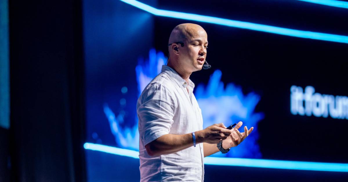 Guilherme Gennari, especialista em biomimética, fala ao público durante o evento IT Forum Trancoso. Ele veste uma camisa branca e utiliza um microfone de cabeça, com um telão ao fundo exibindo o nome do evento em letras brancas (natureza)