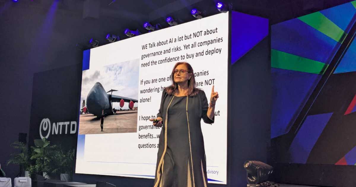 Kay Firth-Butterfield, CEO da Good Tech Advisory, está em pé no palco durante uma apresentação. Ela fala diante de um grande telão que exibe uma imagem de um avião militar e um texto sobre inteligência artificial, governança e riscos. O evento ocorre em um espaço moderno com iluminação de palco e o logotipo da NTT Data visível ao fundo (governança)
