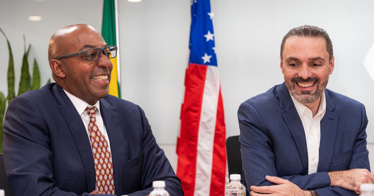 Dois homens bem vestidos, sentados lado a lado numa reunião, sorriem enquanto conversam. Ao fundo, vêem-se as bandeiras do Brasil e dos Estados Unidos, sugerindo um encontro diplomático ou empresarial entre os dois países.