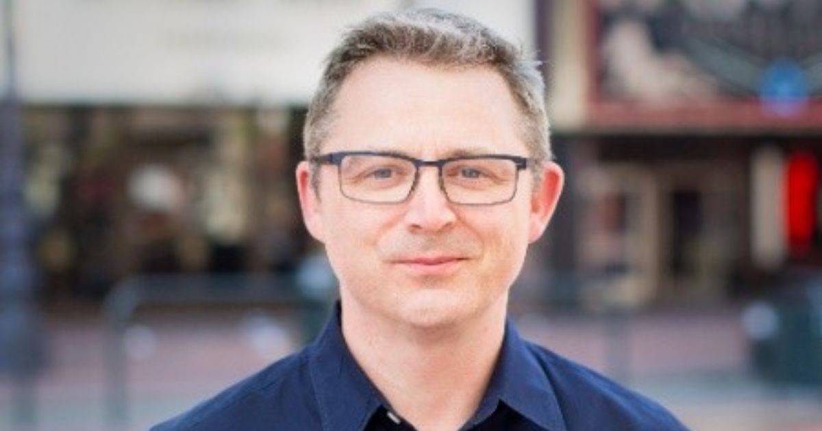 Homem de meia-idade, com cabelo curto e óculos, sorri levemente para a câmara. Está vestido com uma camisa azul-escura e o fundo da imagem mostra um cenário urbano desfocado, sugerindo que a fotografia foi tirada ao ar livre, em contexto citadino