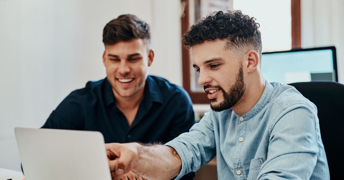 Duas pessoas estão sentadas lado a lado em frente a um laptop. A pessoa à direita faz um gesto com a mão, apontando para a tela do computador, enquanto a outra observa atentamente. As identidades das pessoas foram preservadas com o desfoque dos rostos. Ao fundo, é possível ver uma janela iluminada e um segundo monitor de computador, sugerindo um ambiente de trabalho colaborativo ou reunião informal. (capacitar)
